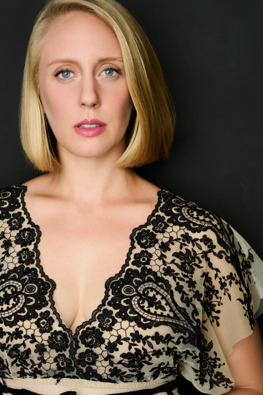 Nicole stares directly at the camera with a serious expression. She wears a black, lacy top with a plunging neckline.